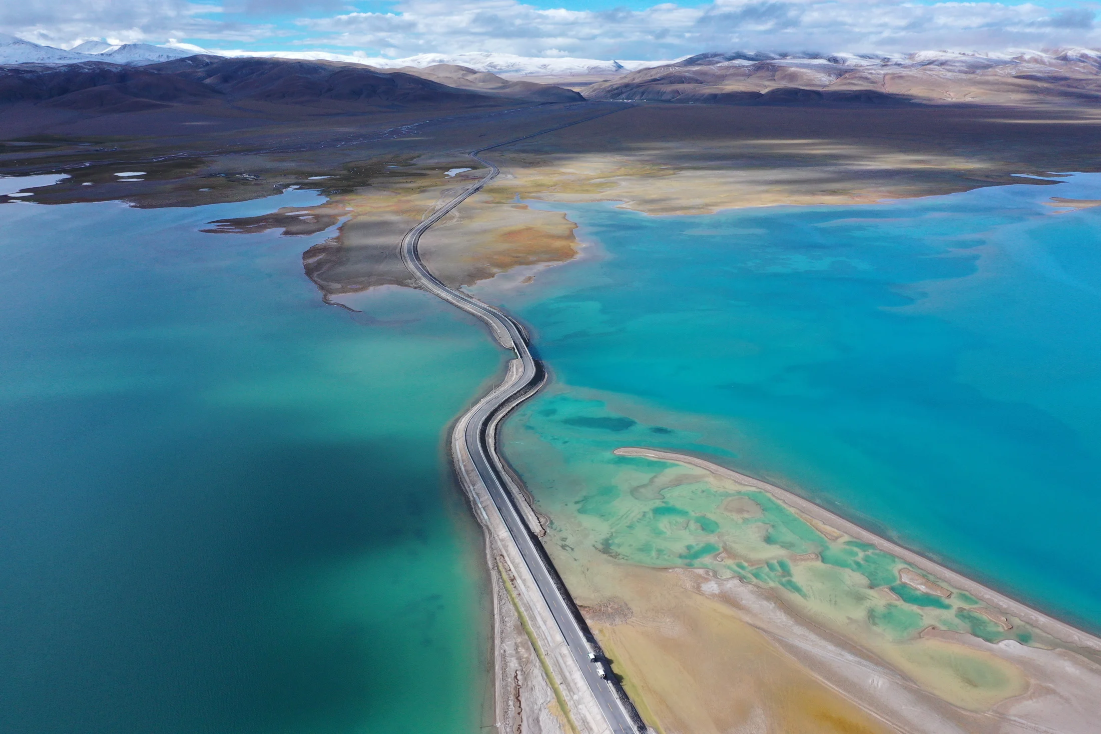 A winding road crosses vivid turquoise water beneath distant mountains